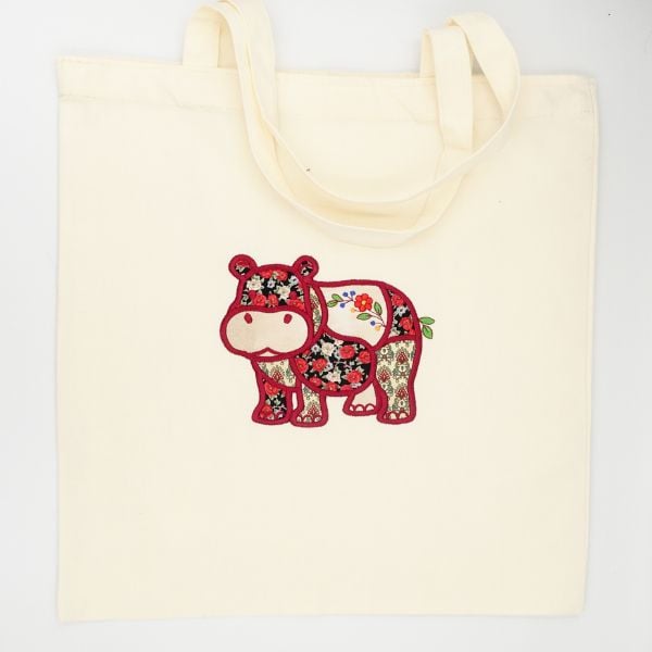 freebie-newsletter-hippo-square | Embroidery Legacy Cream tote bag with a colorful embroidered patchwork hippo design on the front, handles at the top.