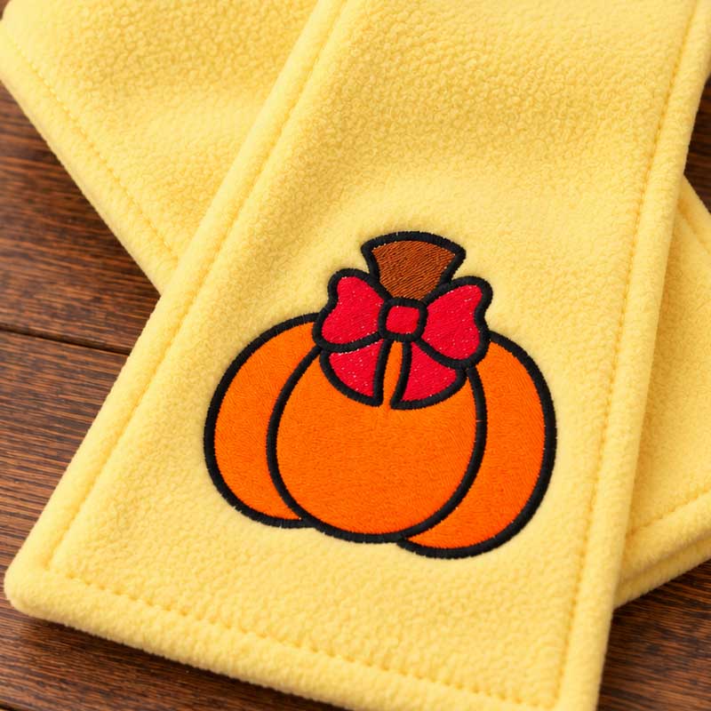 SDCFA2_PumpkinBow_Scarf | Embroidery Legacy Close-up of yellow fleece fabric with an embroidered orange pumpkin and red bow on a wooden surface (decorative item).
